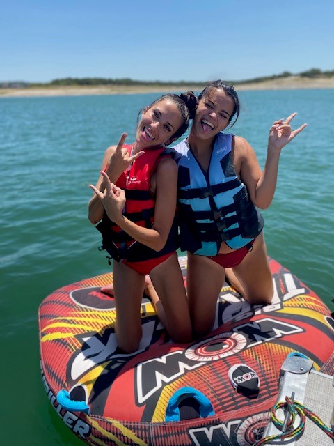 Two teen girls wearing life jackets kneel on a water tube at VIP Marina, Lake Travis, while smiling and making hand gestures
